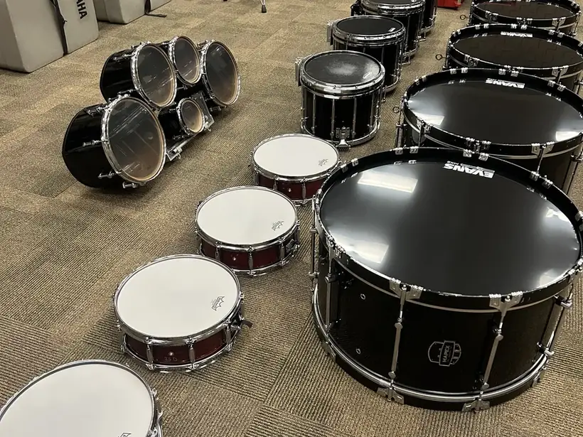 Marching bass drums, marching snares, and concert snares laid out on a band room floor with brand-new heads installed