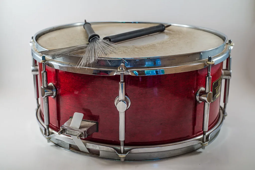 A drum-kit snare drum with wire brushes resting across the batter head